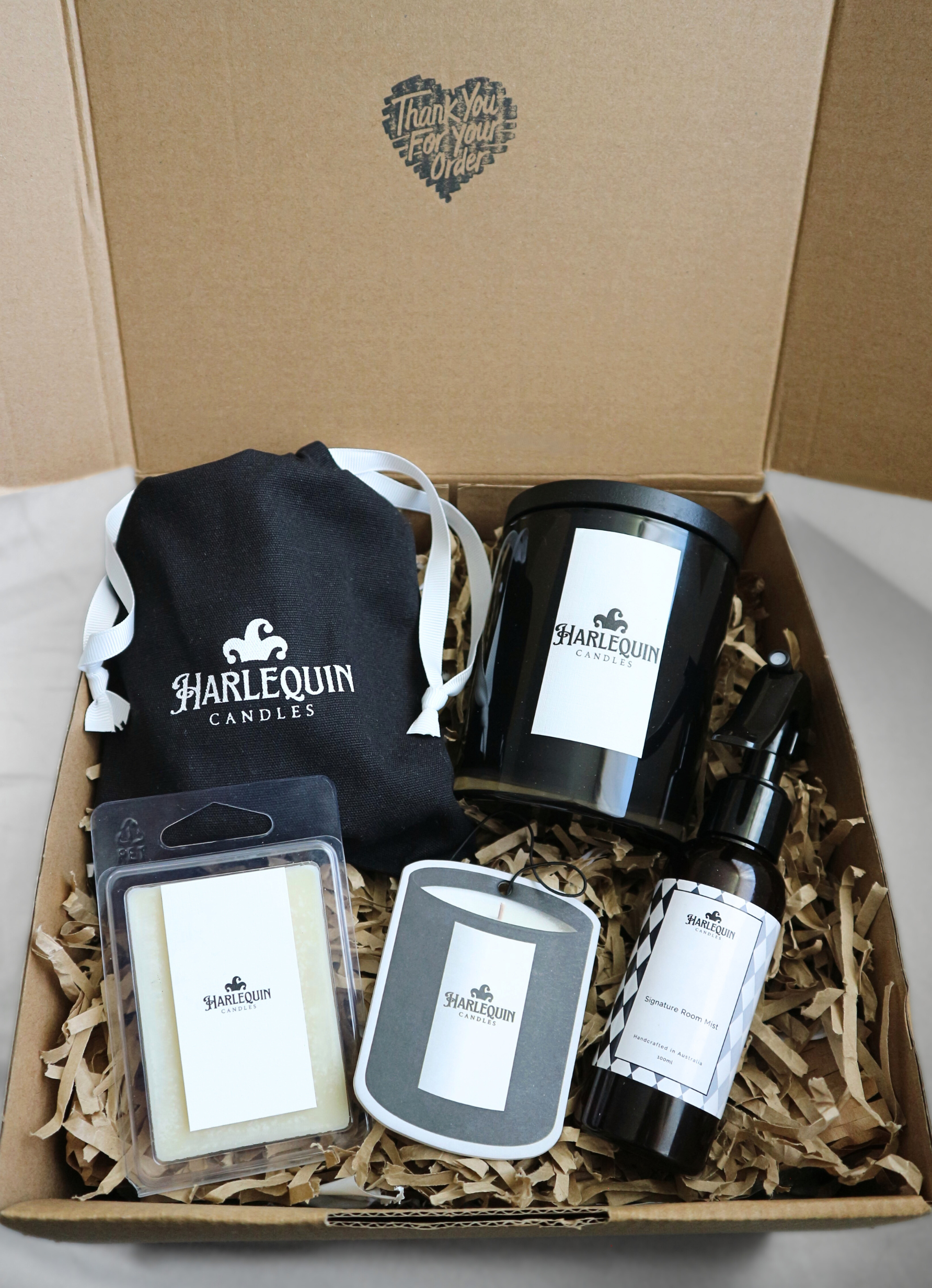 Open Harlequin Candles gift box showcasing a variety of home fragrance items nestled in brown shredded paper. Includes a black jar candle, soy wax melts in a clear clamshell, a room mist spray, and a branded black drawstring bag. 'Thank You For Your Order' is printed on the inside of the box lid. Promotes customizable Australian-made gift sets.