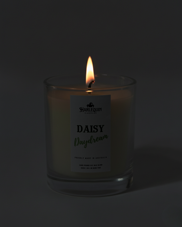Lit Harlequin Candles Daisy Daydream candle in a dark glass jar against a dark background, emphasizing the warm, cozy glow of the flame. Describes a proudly Australian-made, hand-poured soy wax candle.