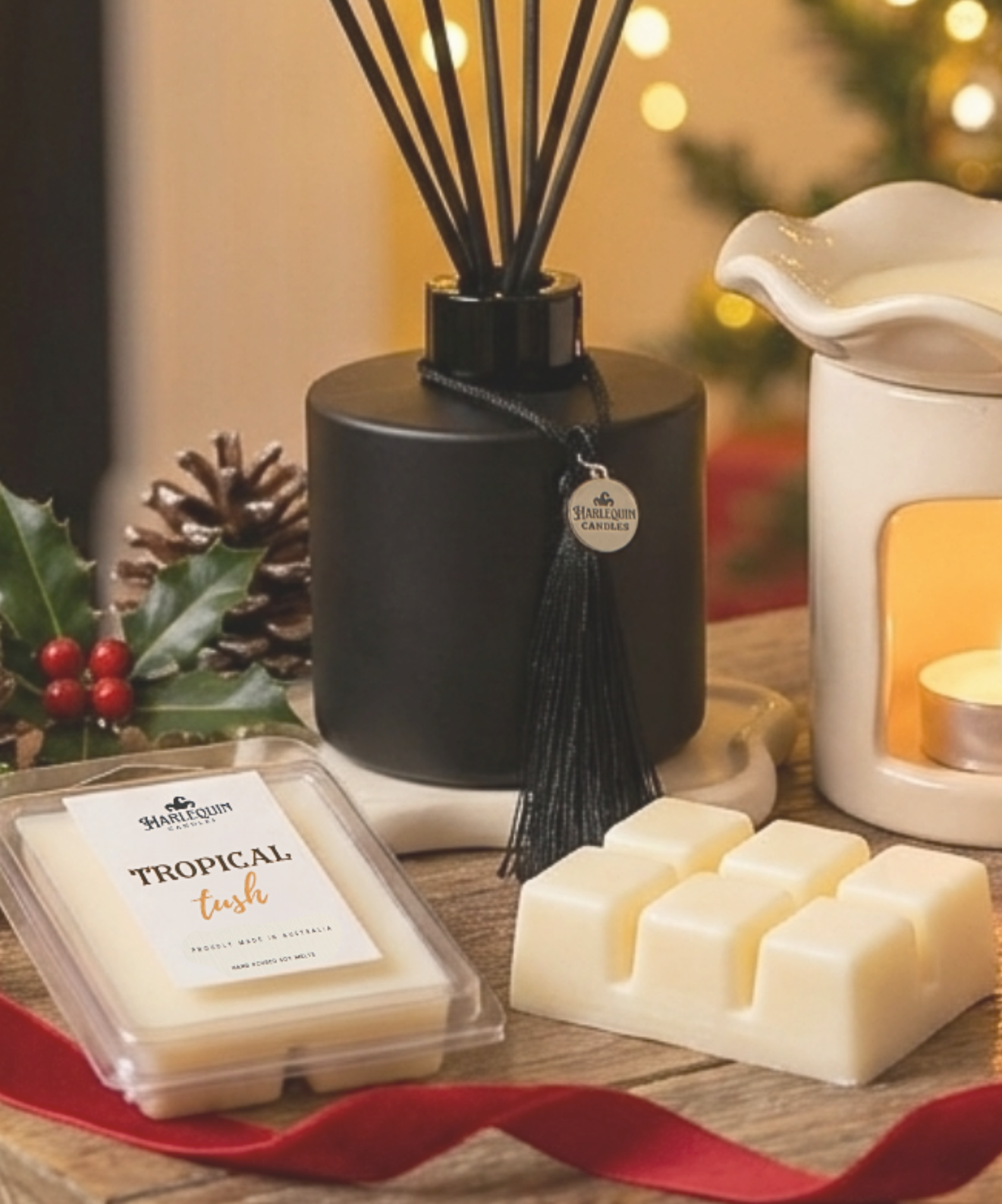 Festive home fragrance display featuring a luxury reed diffuser with black reeds and a stack of heart-shaped soy wax melts on a rustic wooden table. Background features a lit Christmas tree and pinecones. Perfect Australian-made Christmas gift idea.