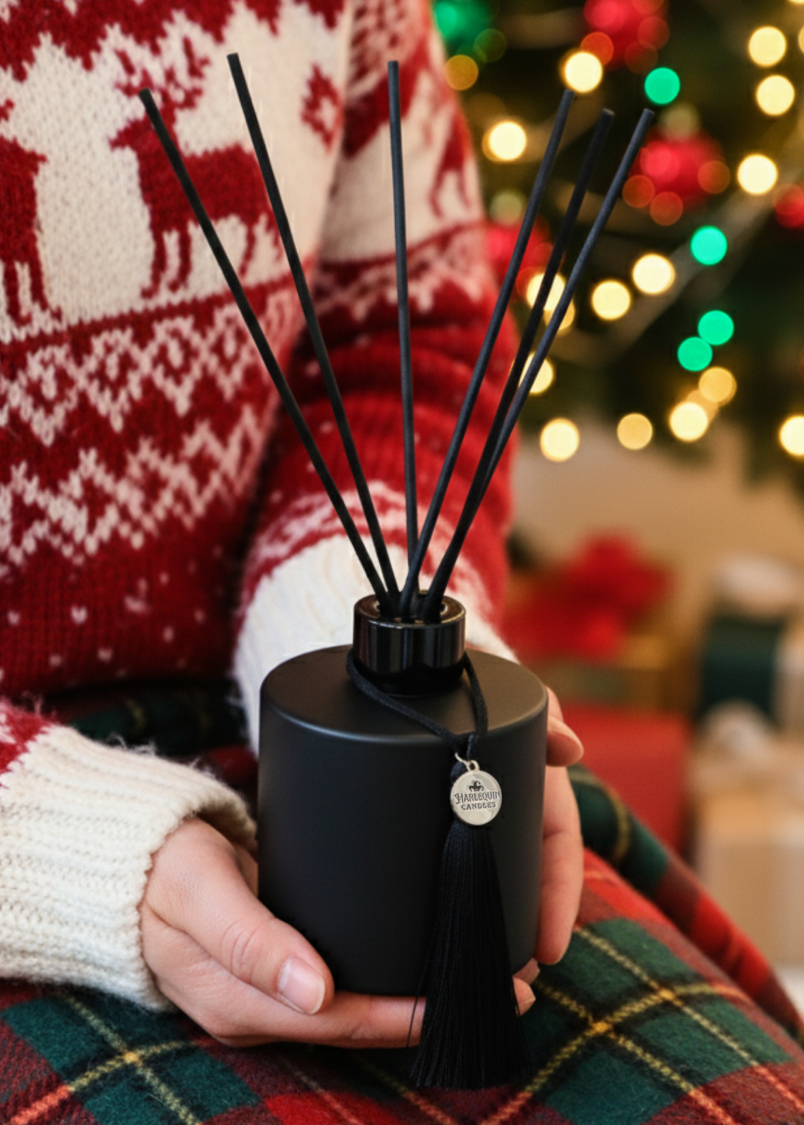 A person in a festive red and white Christmas sweater holding a black Harlequin Candles reed diffuser, featuring a decorative tassel and charm. The background shows a blurred Christmas tree and wrapped gifts, perfectly setting the scene for a handmade Australian Christmas gift or holiday home fragrance.