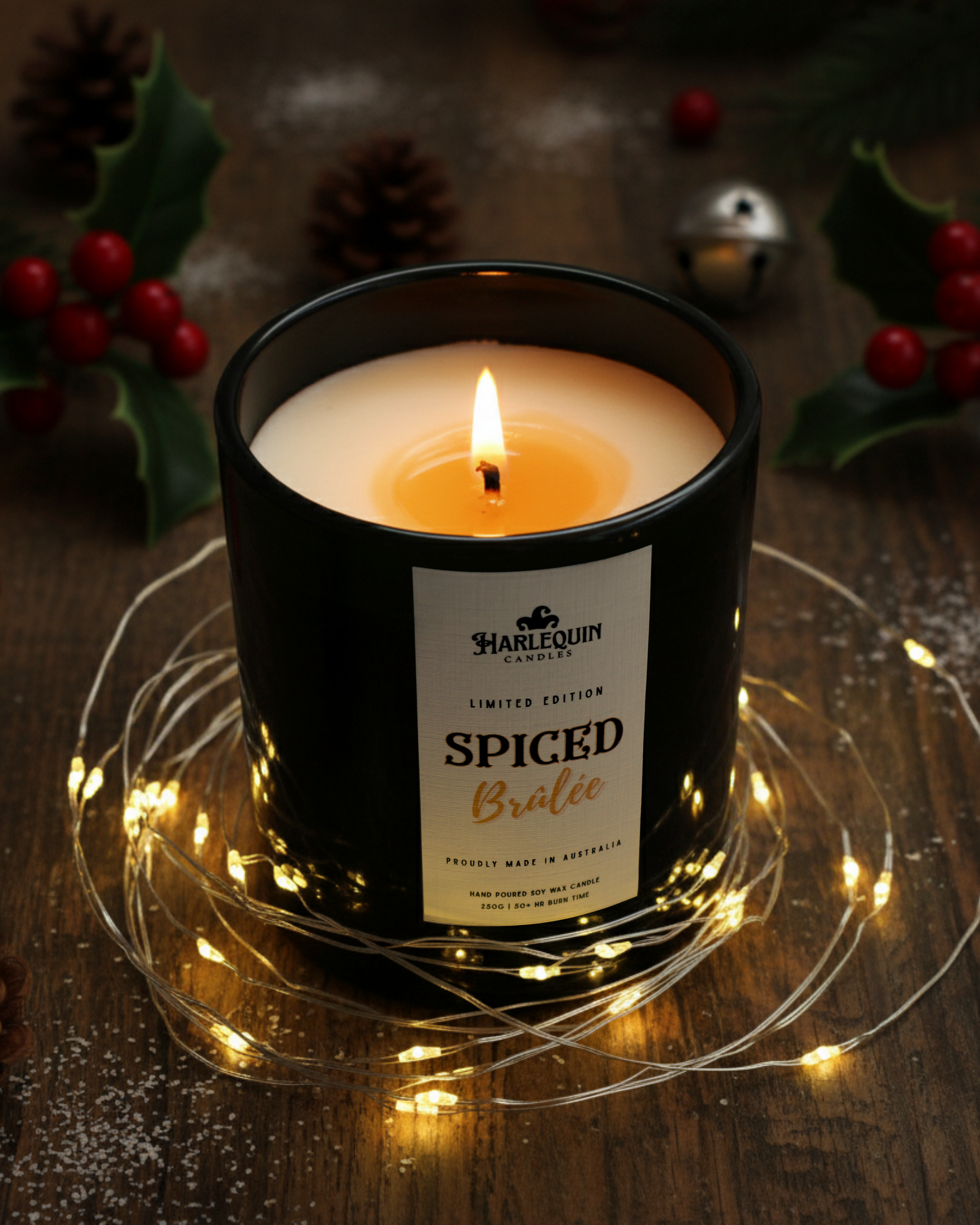 Close-up of a glowing handcrafted Australian soy candle, 'Spiced Brûlée' by Harlequin Candles, featuring festive Christmas decorations like pine needles, red berries, and fairy lights on a dark wood background, emphasizing its limited edition and handmade quality for holiday gifting.