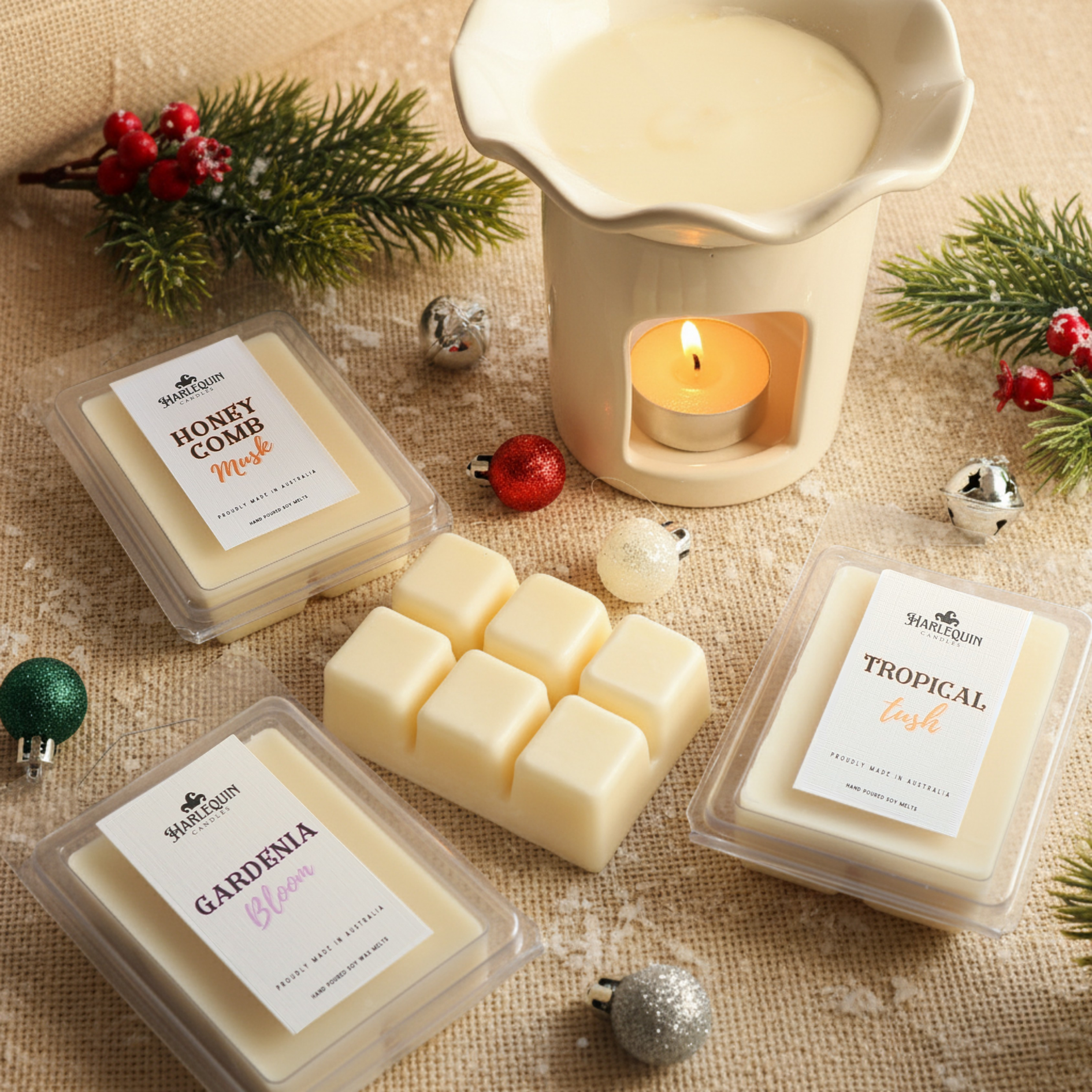 Three packs of Harlequin Candles handmade Australian-made soy wax melts in Honeycomb Musk, Gardenia Bloom, and Tropical Tush scents, arranged with a lit wax warmer and festive Christmas ornaments on a burlap background, perfect for stocking stuffers and unique Australian Christmas gifts.