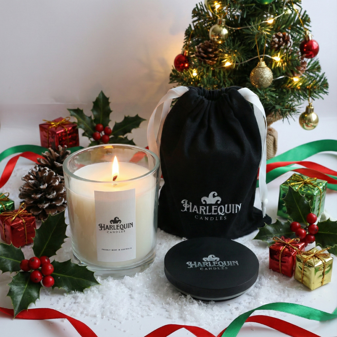 Lit handmade soy candle by Harlequin Candles displayed in fake snow with a luxury black gift bag, Christmas tree, and festive ribbons. An elegant, Australian-made home fragrance gift perfect for Christmas.
