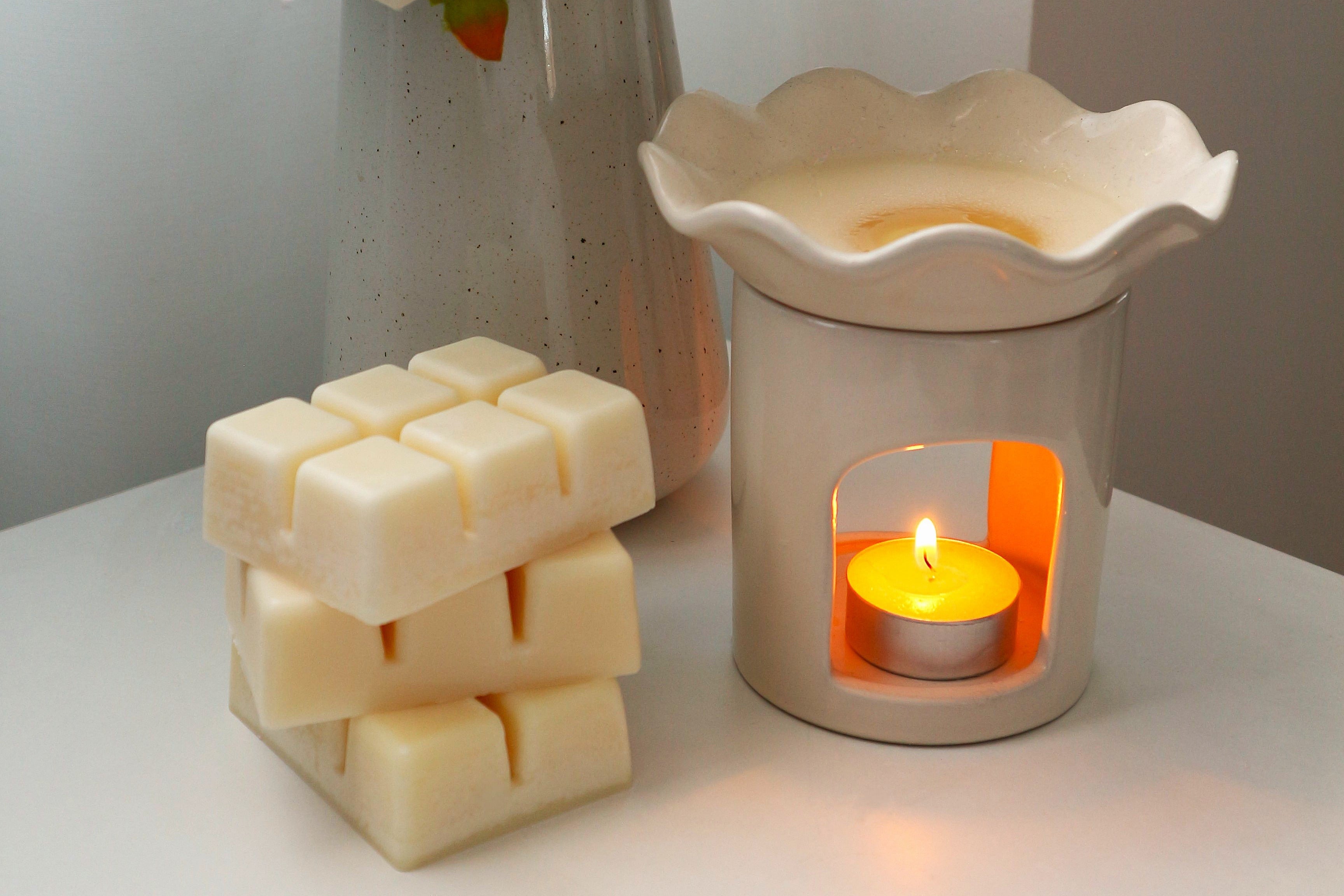 A stack of natural Harlequin Candles 100% soy wax melts next to a white ceramic wax warmer with a lit tealight and melted wax, on a white surface with a vase of white flowers in the background. Showcases eco-friendly home fragrance.