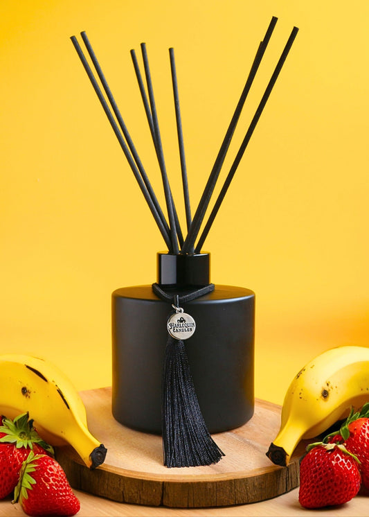 Harlequin Candles Banana Drama reed diffuser in a matte black bottle with black reeds and a decorative black tassel with a silver logo charm, displayed on a wooden slice with fresh bananas and strawberries against a bright yellow background.