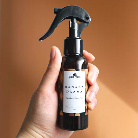 A Harlequin Candles Banana Drama Signature Room Mist in an amber spray bottle with a black trigger cap, centered on a two-toned background of light yellow and white. The label clearly shows 'Harlequin Candles', 'Banana Drama', 'Signature Room Mist', and 'Handcrafted in Australia', highlighting this vibrant Australian-made home spray.