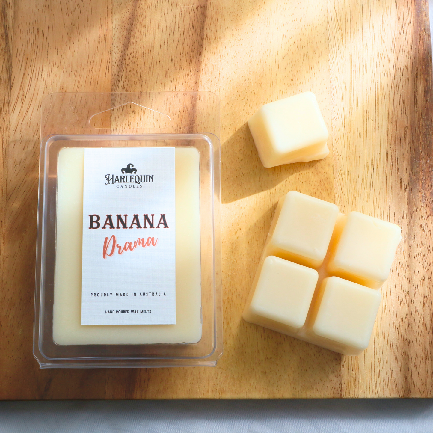 Harlequin Candles Banana Drama soy wax melts, displayed in clear plastic clamshell packaging with the brand label. The label reads 'BANANA Drama' and 'PROUDLY MADE IN AUSTRALIA HAND POURED WAX MELTS'. Several individual wax melt cubes are scattered on a light wooden cutting board, showcasing the fruity home fragrance product and its local origin.
