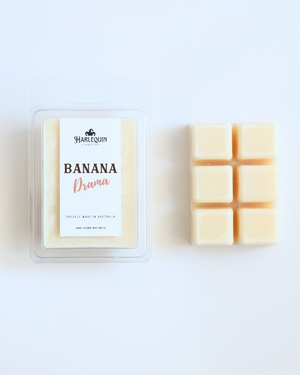 Harlequin Candles Banana Drama soy wax melts, displayed in clear plastic clamshell packaging with the brand label. The label reads 'BANANA Drama' and 'PROUDLY MADE IN AUSTRALIA HAND POURED WAX MELTS'. Several individual wax melt cubes are scattered on a light wooden cutting board, showcasing the fruity home fragrance product and its local origin.