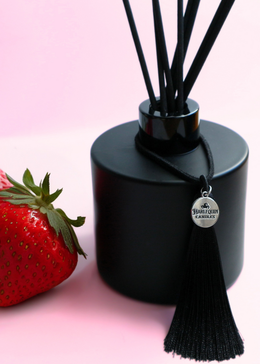 Harlequin Candles Berry Bubbly (Strawberries & Champagne) reed diffuser in a matte black bottle with black reeds and a decorative black tassel with a silver logo charm. A vibrant red rose lies beside the diffuser on a pink background, evoking a celebratory and fruity home fragrance.