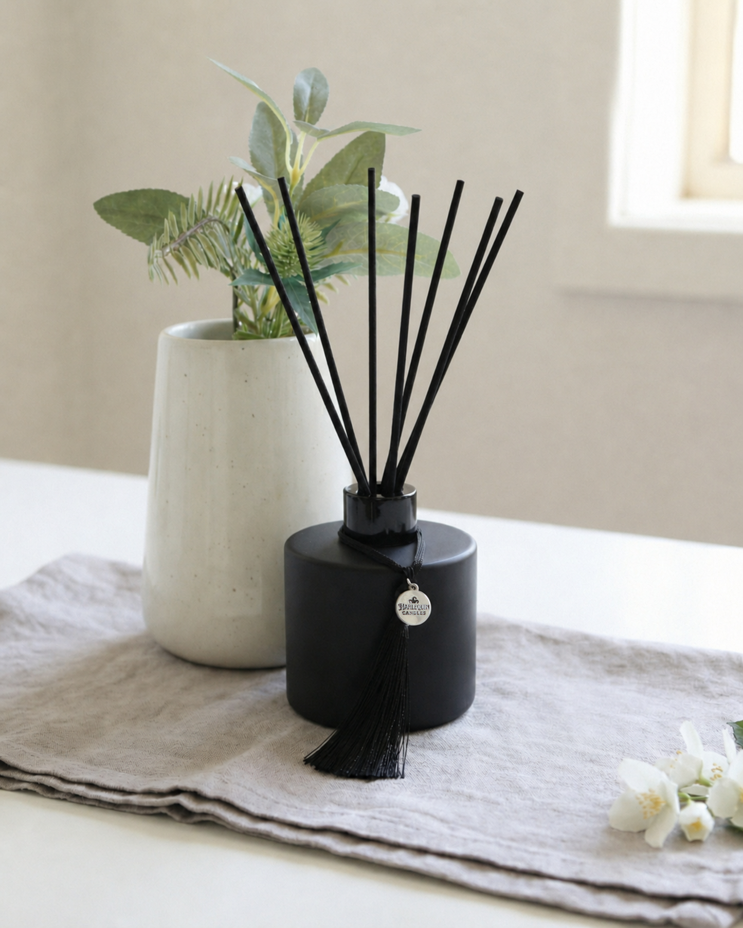 Harlequin Candles black reed diffuser with black reeds and tassel on linen cloth beside white flowers and ceramic vase, hand-poured in Melbourne