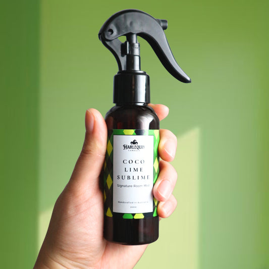 A Harlequin Candles Coco Lime Sublime Signature Room Mist in an amber spray bottle with a black trigger cap, placed against a two-toned background of light green and white. The label clearly shows 'Harlequin Candles', 'Coco Lime Sublime', 'Signature Room Mist', and 'Handcrafted in Australia', highlighting this tropical Australian-made home spray.