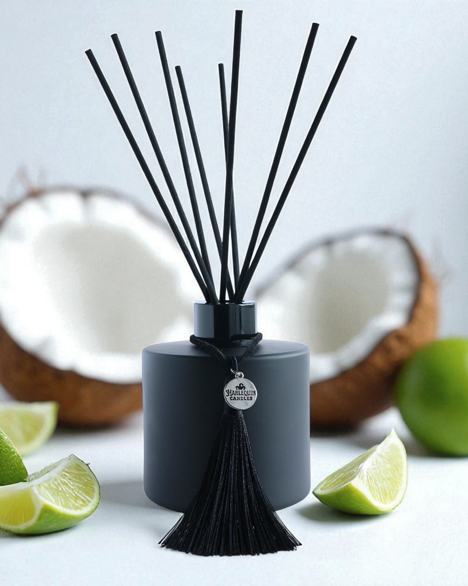 Harlequin Candles Mint Mojito reed diffuser in a matte black bottle with black reeds and a decorative black tassel with a silver logo charm, surrounded by fresh lime wedges and fresh cut coconut.
