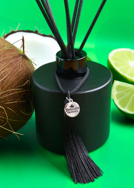 Harlequin Candles Coconut Lime reed diffuser in a matte black bottle with black reeds and a decorative black tassel with a silver logo charm, surrounded by fresh coconut and lime halves on a vibrant green background.