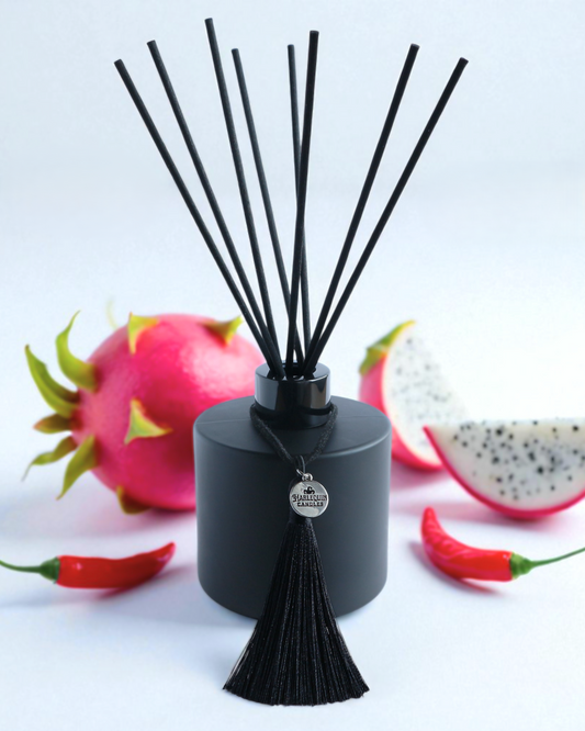 Harlequin Candles Fruit & Fire (Dragonfruit & Chipotle) reed diffuser in a matte black bottle with black reeds and a decorative black tassel with a silver logo charm. Two vibrant pink roses flank the diffuser, set against a bold red background, evoking a passionate and exotic home fragrance.