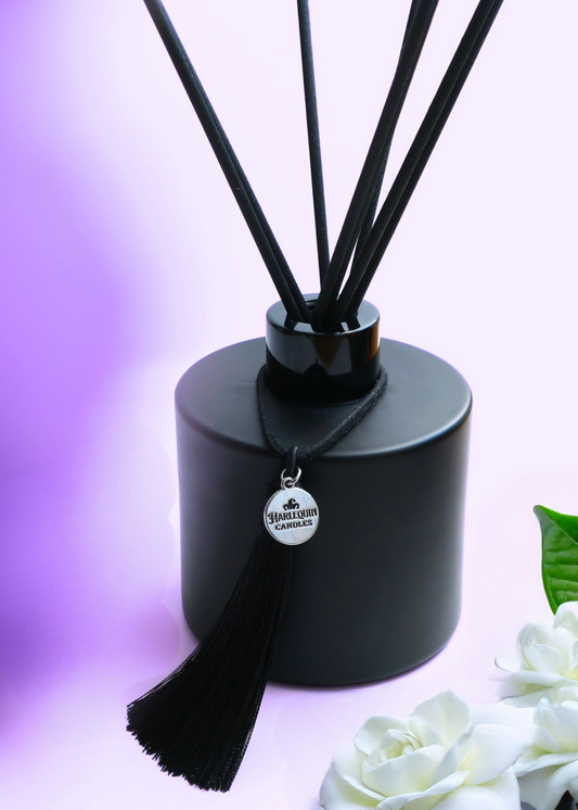 Harlequin Candles Gardenia Bloom reed diffuser in a matte black bottle with black reeds and a decorative black tassel with a silver logo charm. A large, pristine white gardenia flower is next to the diffuser, set against a soft purple background, evoking a lush and elegant home fragrance.