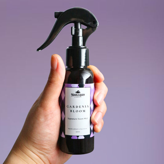A Harlequin Candles Gardenia Bloom Signature Room Mist in an amber spray bottle with a black trigger cap, placed against a two-toned background of light pink and white. The label clearly shows 'Harlequin Candles', 'Gardenia Bloom', 'Signature Room Mist', and 'Handcrafted in Australia', highlighting this lush floral Australian-made home spray.