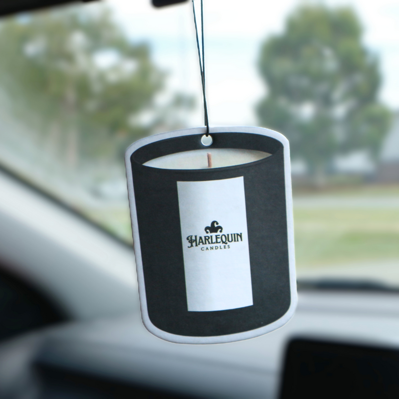A Harlequin Candles candle-shaped hanging car freshener with a white label prominently featuring the brand logo, suspended from a car's rearview mirror. The interior of the car is visible, with blurred trees and buildings through the windshield, showcasing a stylish Australian car air freshener.