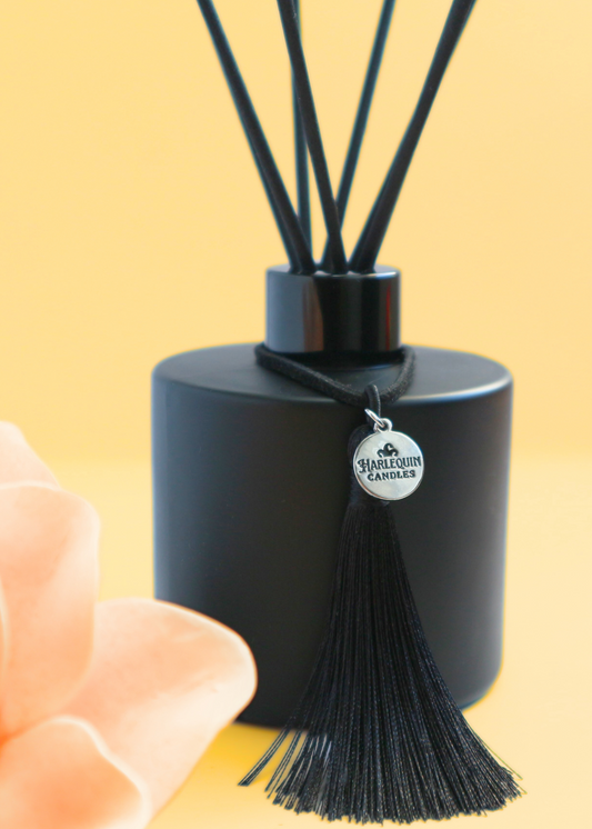 Harlequin Candles Honeycomb Musk reed diffuser in a matte black bottle with black reeds and a decorative black tassel with a silver logo charm. A soft orange-toned flower is blurred in the foreground, set against a warm yellow background, evoking an inviting and comforting home aroma.