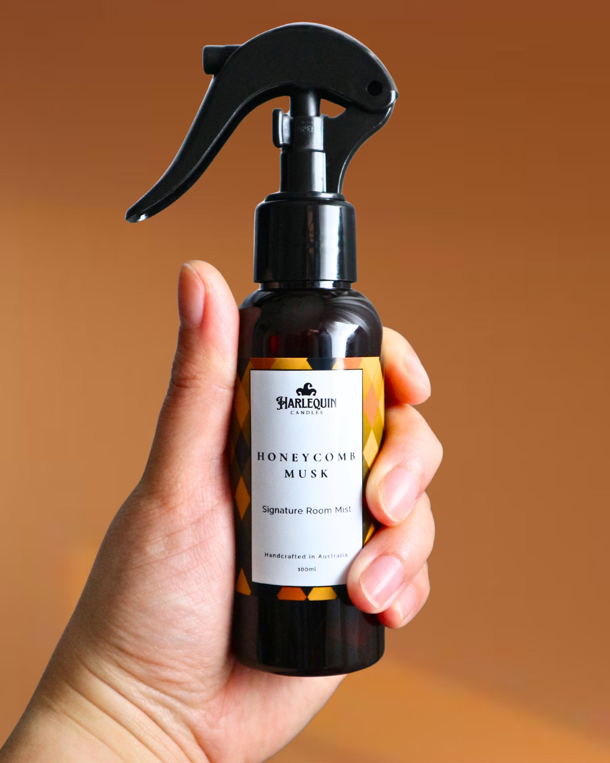 A Harlequin Candles Honeycomb Musk Signature Room Mist in an amber spray bottle with a black trigger cap, placed against a two-toned background of light yellow and white. The label clearly shows 'Harlequin Candles', 'Honeycomb Musk', 'Signature Room Mist', and 'Handcrafted in Australia', highlighting this warm and comforting Australian-made home spray.