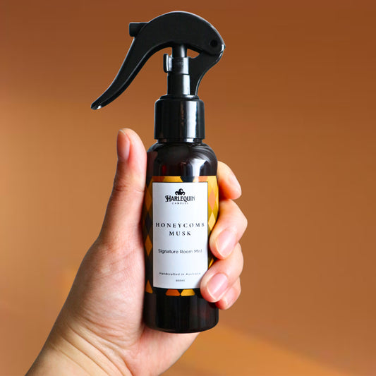 A Harlequin Candles Honeycomb Musk Signature Room Mist in an amber spray bottle with a black trigger cap, placed against a two-toned background of light yellow and white. The label clearly shows 'Harlequin Candles', 'Honeycomb Musk', 'Signature Room Mist', and 'Handcrafted in Australia', highlighting this warm and comforting Australian-made home spray.
