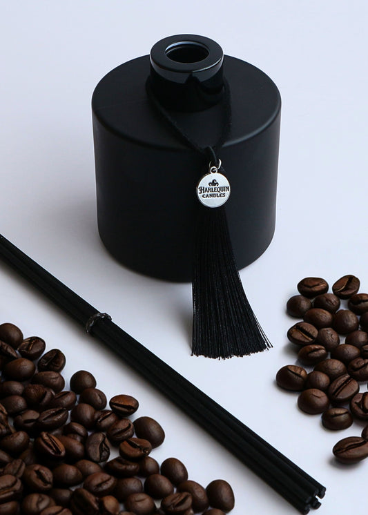 Harlequin Candles Italian Espresso reed diffuser in a matte black bottle with a decorative black tassel and silver logo charm, surrounded by roasted coffee beans and a bundle of black reeds, on a light grey background.