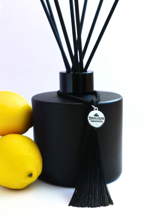 Harlequin Candles Lemon Liquor (Limoncello scented) reed diffuser in a matte black bottle with black reeds and a decorative black tassel with a silver logo charm, positioned alongside two fresh lemons on a white background.