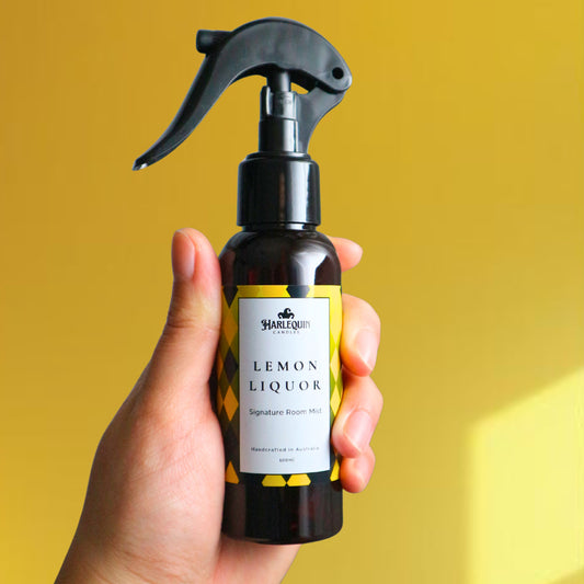 A Harlequin Candles Lemon Liquor (Limoncello scented) Signature Room Mist in an amber spray bottle with a black trigger cap, placed against a two-toned background of light yellow and white. The label clearly shows 'Harlequin Candles', 'Lemon Liquor', 'Signature Room Mist', and 'Handcrafted in Australia', highlighting this zesty Australian-made home spray.