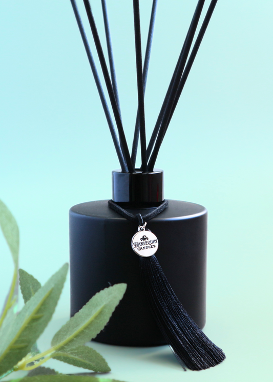 Harlequin Candles Lemon Myrtle reed diffuser in a matte black bottle with black reeds and a decorative black tassel with a silver logo charm. A fresh green leafy branch is in the foreground, set against a serene light blue-green background, emphasizing this Australian botanical home fragrance.