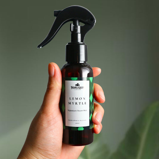 A Harlequin Candles Lemon Myrtle Signature Room Mist in an amber spray bottle with a black trigger cap, placed against a two-toned background of light green and white. The label clearly shows 'Harlequin Candles', 'Lemon Myrtle', 'Signature Room Mist', and 'Handcrafted in Australia', highlighting this refreshing Australian-made home spray.
