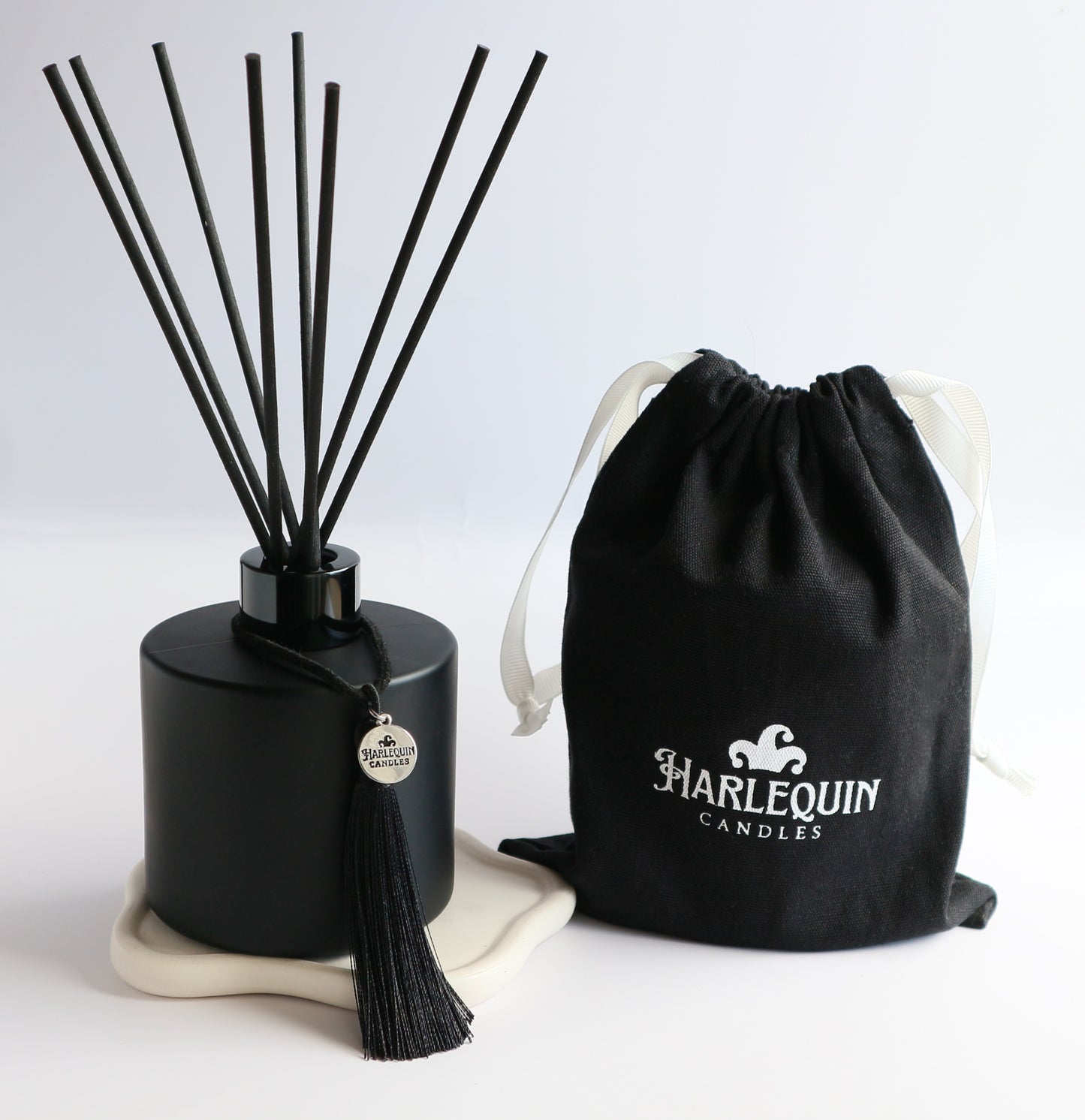 A sleek matte black Harlequin Candles reed diffuser bottle with black reeds, a black tassel charm, and a black drawstring gift bag featuring the white Harlequin Candles logo. The items are artfully arranged on a subtle white coaster against a clean white background, showcasing a luxury home fragrance product.