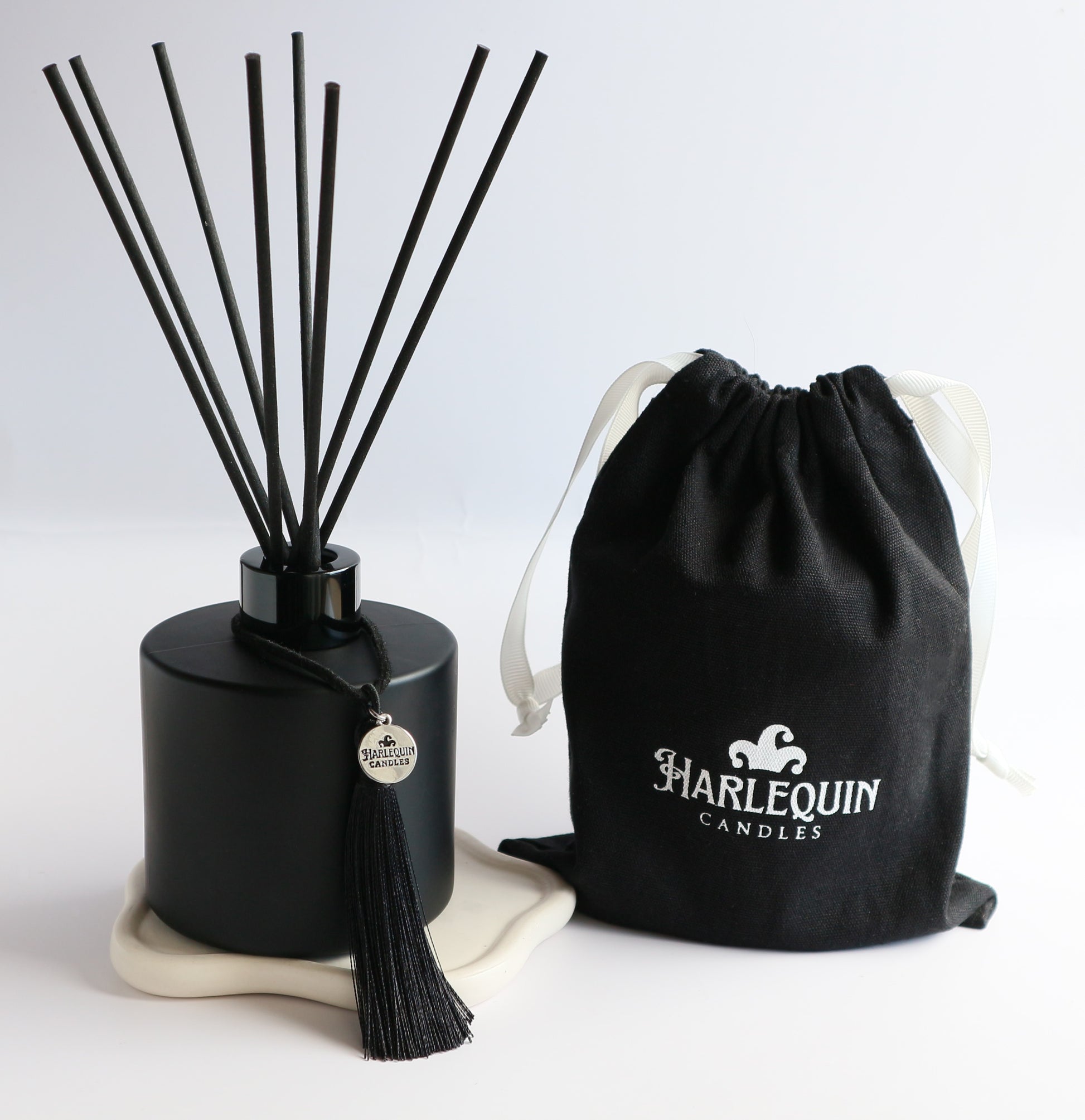 A sleek matte black Harlequin Candles reed diffuser bottle with black reeds, a black tassel charm, and a black drawstring gift bag featuring the white Harlequin Candles logo. The items are artfully arranged on a subtle white coaster against a clean white background, showcasing a luxury home fragrance product.