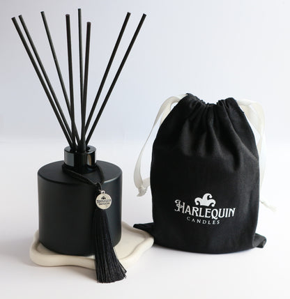 A sleek matte black Harlequin Candles reed diffuser bottle with black reeds, a black tassel charm, and a black drawstring gift bag featuring the white Harlequin Candles logo. The items are artfully arranged on a subtle white coaster against a clean white background, showcasing a luxury home fragrance product.
