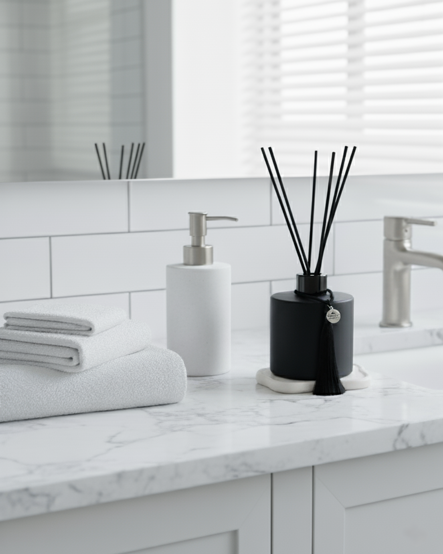 A Harlequin Candles black reed diffuser with a tassel adding a touch of luxury to a serene, white bathroom setting.