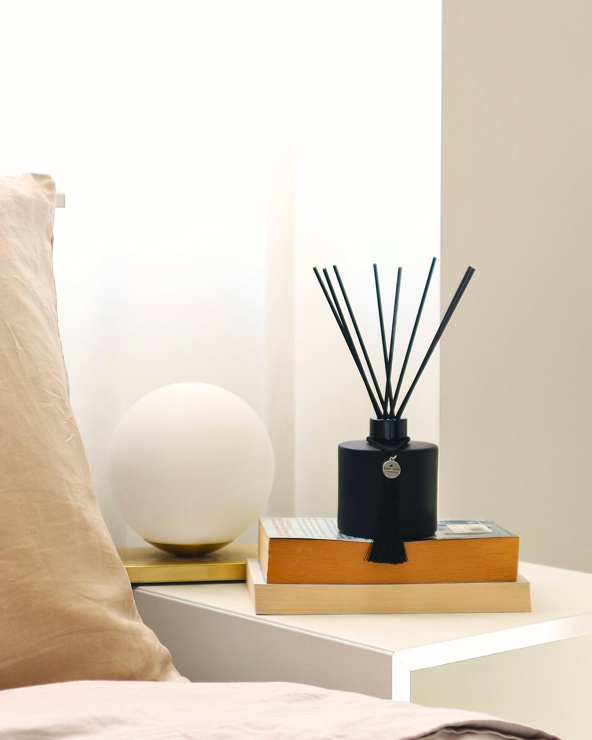 Harlequin Candles matte black reed diffuser with black reeds and a decorative tassel, elegantly displayed on a stack of books on a bedside table in a modern bedroom. A spherical white lamp and neutral-toned pillow complete the serene home fragrance setting.
