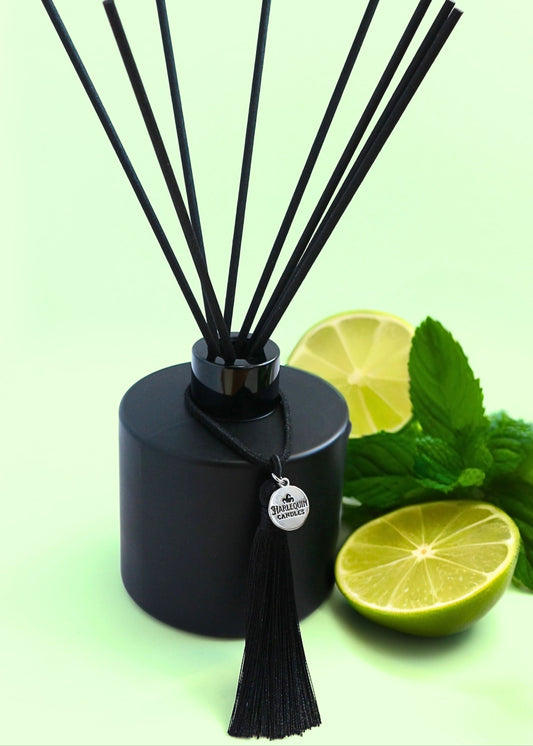 Harlequin Candles Mint Mojito reed diffuser in a matte black bottle with black reeds and a decorative black tassel with a silver logo charm, surrounded by fresh lime slices and mint leaves on a light green background.