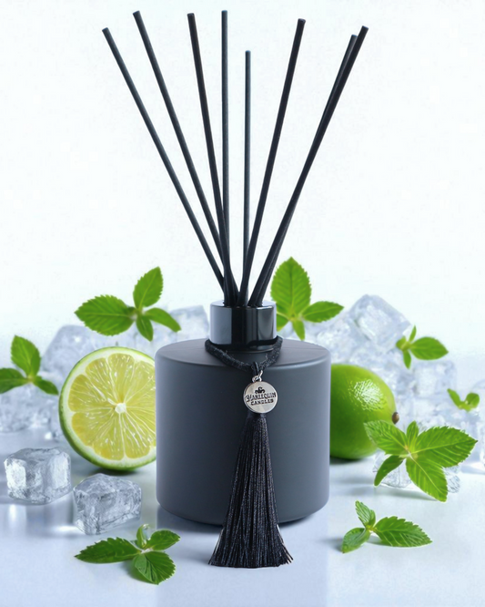 Harlequin Candles Mint Mojito reed diffuser in a matte black bottle with black reeds and a decorative black tassel with a silver logo charm, surrounded by fresh lime slices and mint leaves on a light green background.
