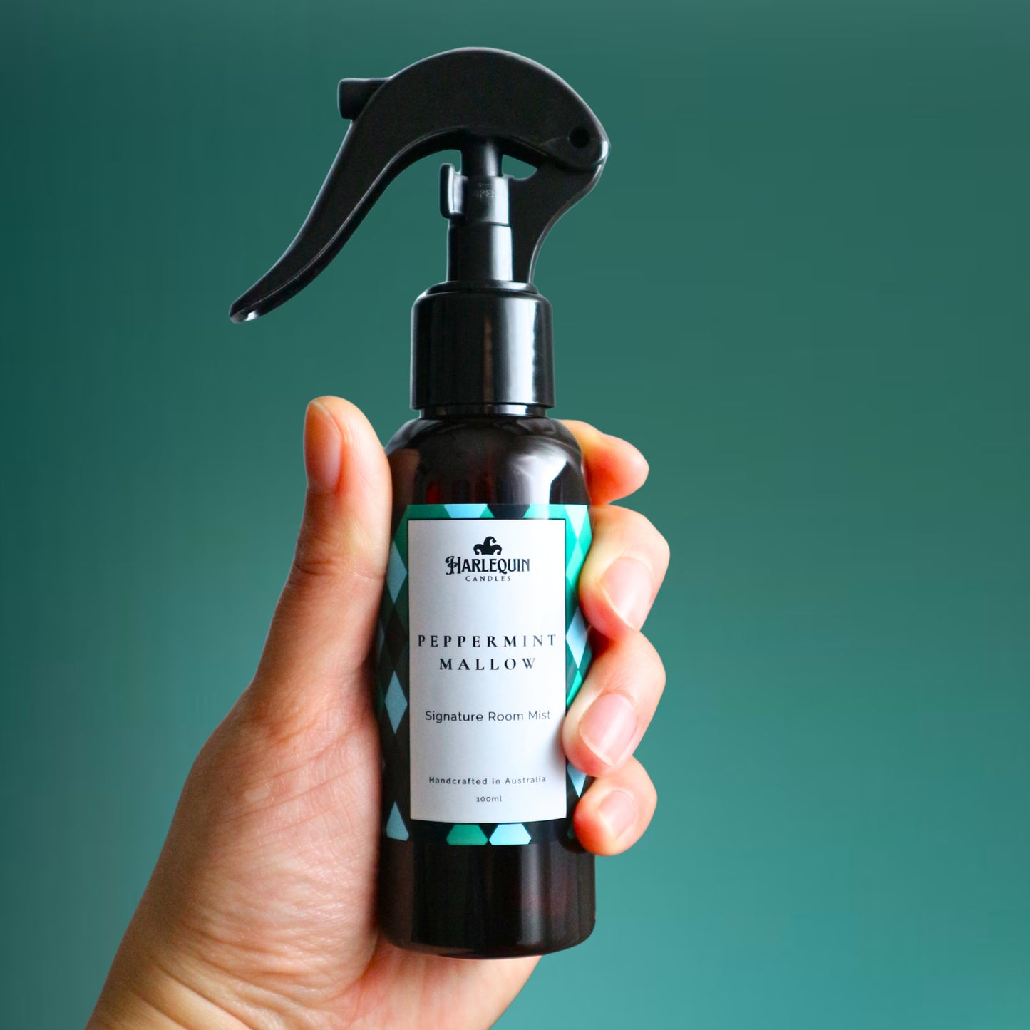 A Harlequin Candles Peppermint Mallow Signature Room Mist in an amber spray bottle with a black trigger cap, placed against a two-toned background of light blue and white. The label clearly shows 'Harlequin Candles', 'Peppermint Mallow', 'Signature Room Mist', and 'Handcrafted in Australia', highlighting this sweet mint Australian-made home spray.