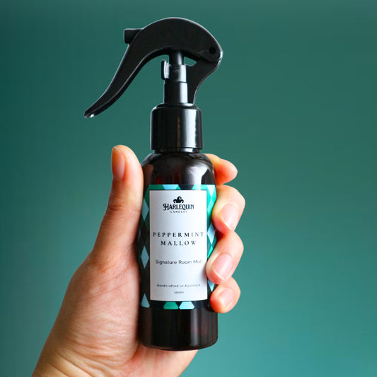 A Harlequin Candles Peppermint Mallow Signature Room Mist in an amber spray bottle with a black trigger cap, placed against a two-toned background of light blue and white. The label clearly shows 'Harlequin Candles', 'Peppermint Mallow', 'Signature Room Mist', and 'Handcrafted in Australia', highlighting this sweet mint Australian-made home spray.