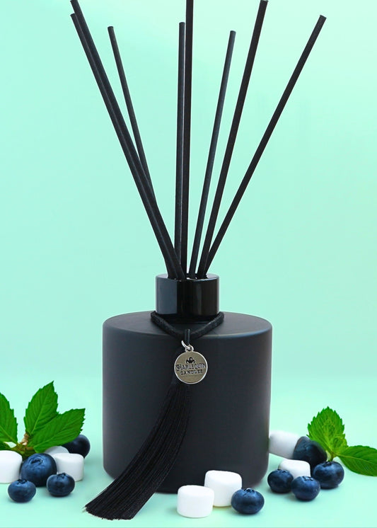 Harlequin Candles Peppermint Marshmallow reed diffuser in a matte black bottle with black reeds and a decorative black tassel with a silver logo charm, surrounded by fresh blueberries, mint leaves, and white marshmallows on a light green background.