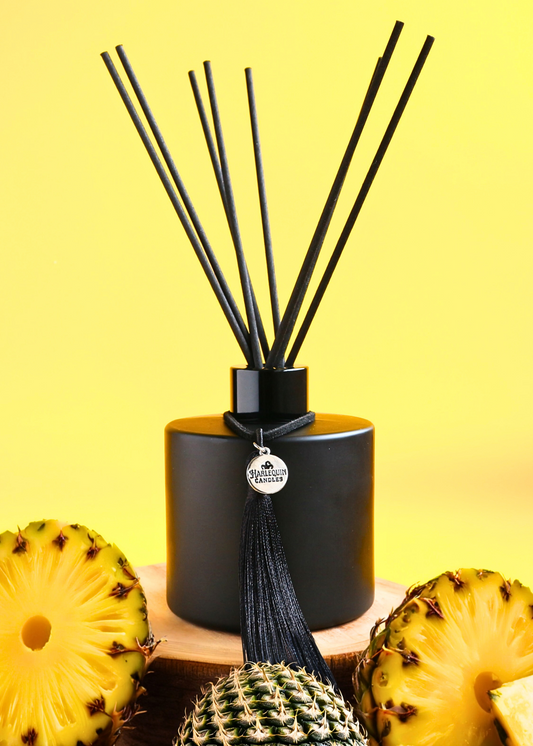 Harlequin Candles Piña Perfection (Pina Colada scent) reed diffuser in a matte black bottle with black reeds and a decorative black tassel, on a wooden surface with sliced pineapple against a yellow background.