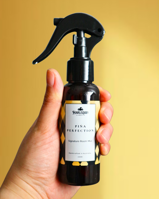 A Harlequin Candles Pina Colada scented Signature Room Mist in an amber spray bottle with a black trigger cap, placed against a two-toned background of light yellow and white. The label clearly shows 'Harlequin Candles', 'Pina Perfection', 'Signature Room Mist', and 'Handcrafted in Australia', highlighting this elegant Australian-made home spray.
