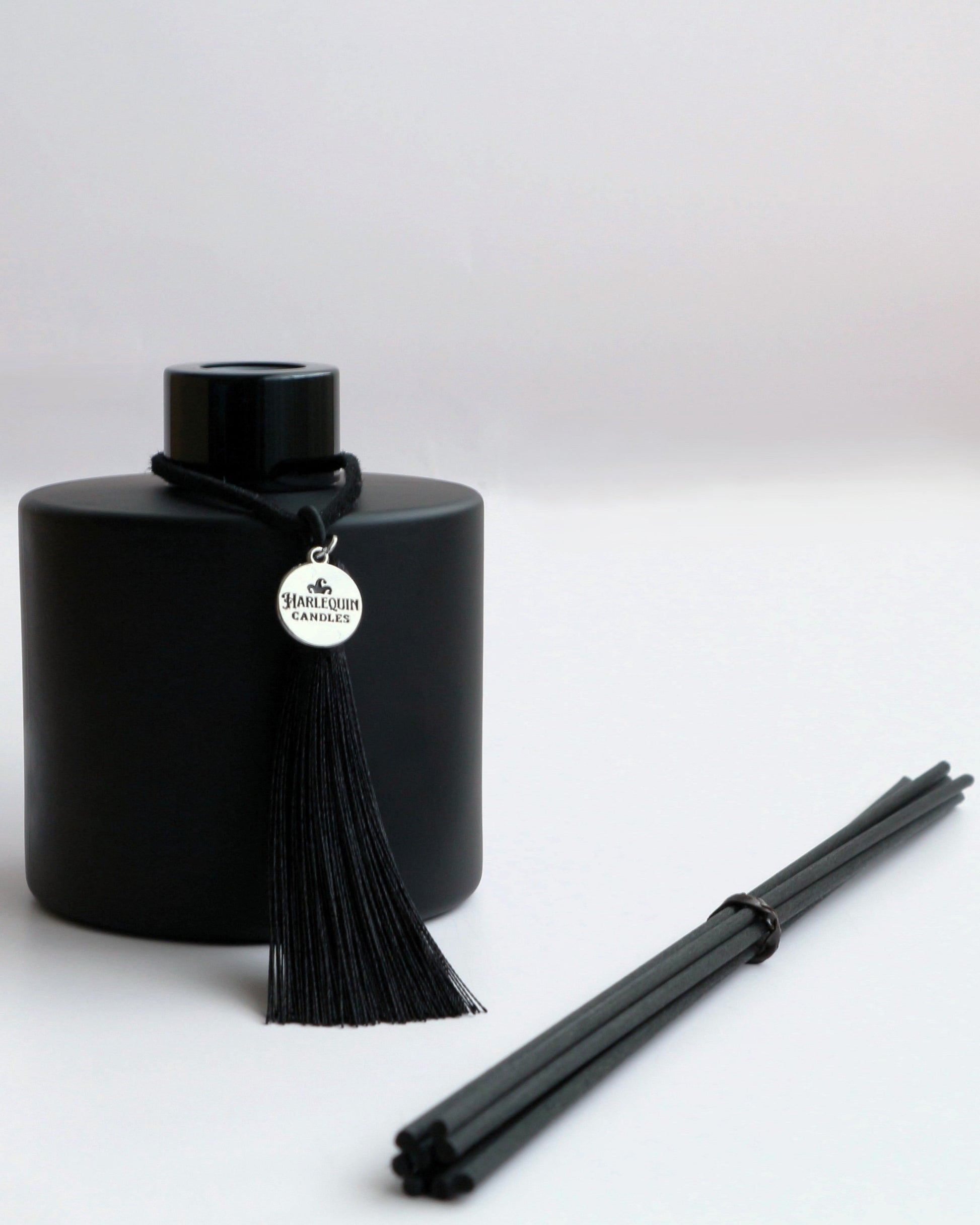A Harlequin Candles premium matte black reed diffuser bottle with a black tassel and silver logo charm, displayed next to a bundle of black diffuser reeds, all on a clean white background. Showcases a stylish home fragrance diffuser set.
