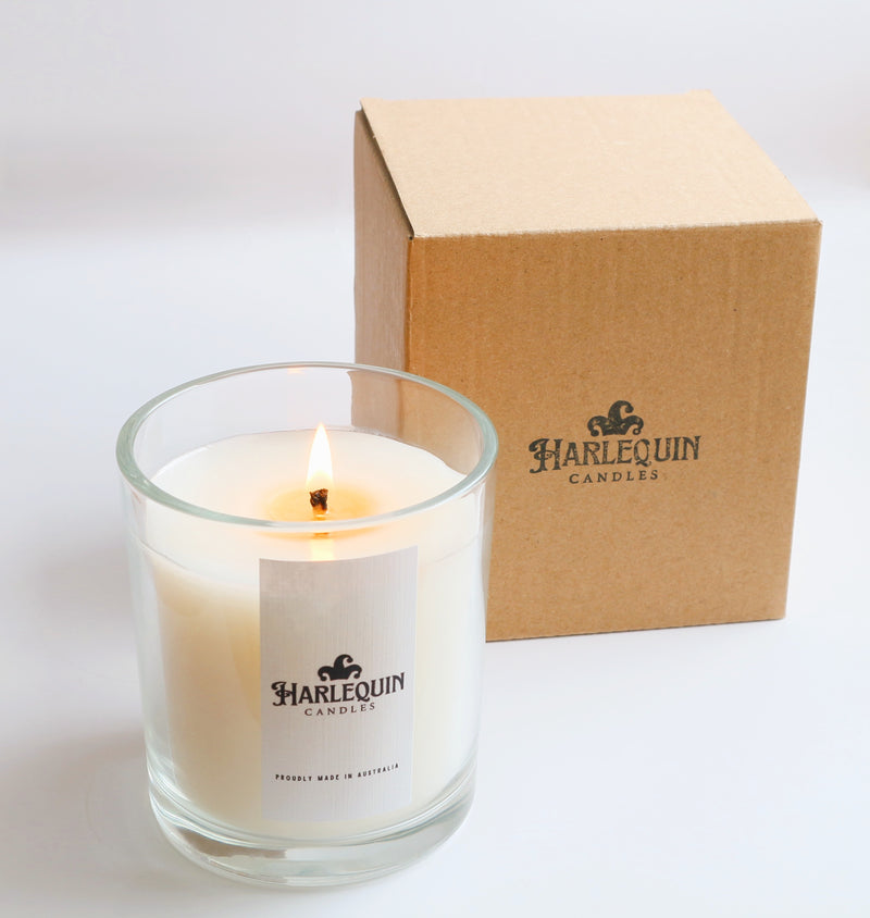 A lit Harlequin Candles premium soy candle in a clear glass jar with a white label prominently displaying the brand logo and 'PROUDLY MADE IN AUSTRALIA' text. Next to it, a plain cardboard box with the Harlequin Candles logo. All are displayed on a white background, highlighting quality Australian craftsmanship.