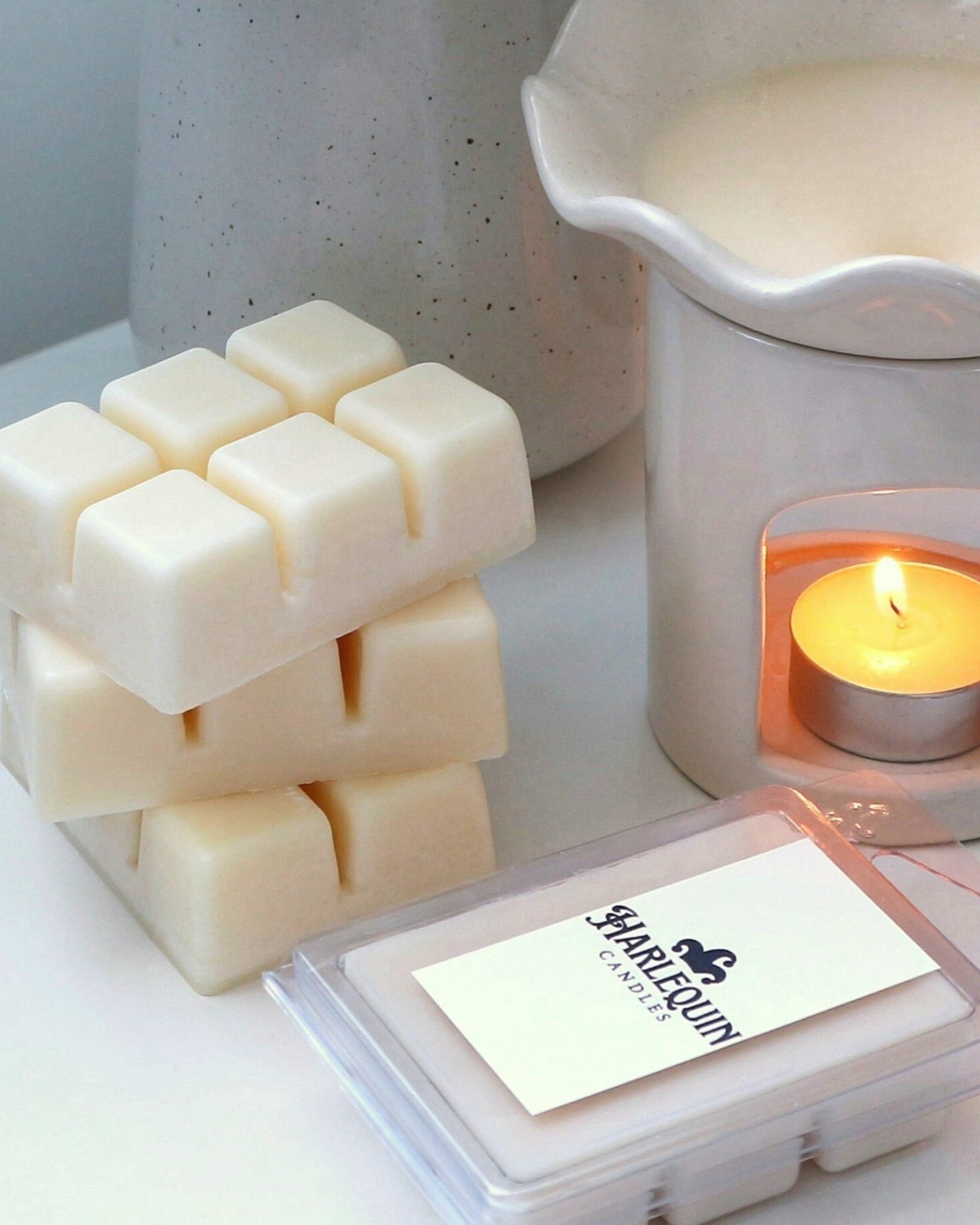 A stack of Harlequin Candles premium soy wax melts next to a white ceramic wax warmer with a lit tealight candle and melted wax. A clear plastic clamshell of wax melts with the Harlequin Candles label is in the foreground, showcasing eco-friendly home fragrance.