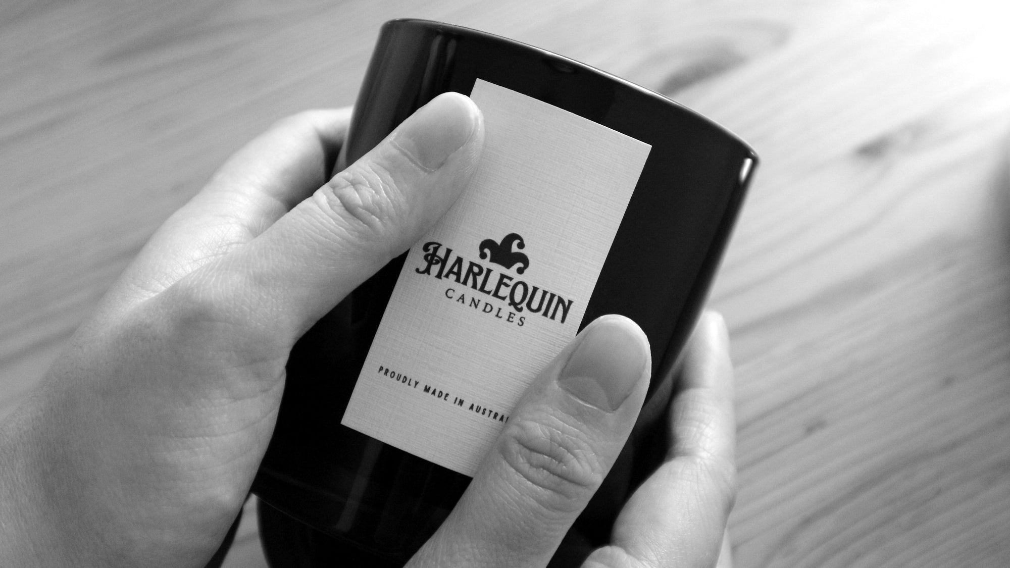Close-up of hands gently holding a black Harlequin Candles product, clearly showing the white label with the brand logo and 'PROUDLY MADE IN AUSTRALIA' text. Highlights the local craftsmanship of Harlequin Candles.