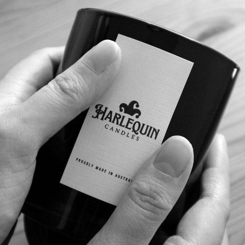 Close-up of hands gently holding a black Harlequin Candles product, clearly showing the white label with the brand logo and 'PROUDLY MADE IN AUSTRALIA' text. Highlights the local craftsmanship of Harlequin Candles.
