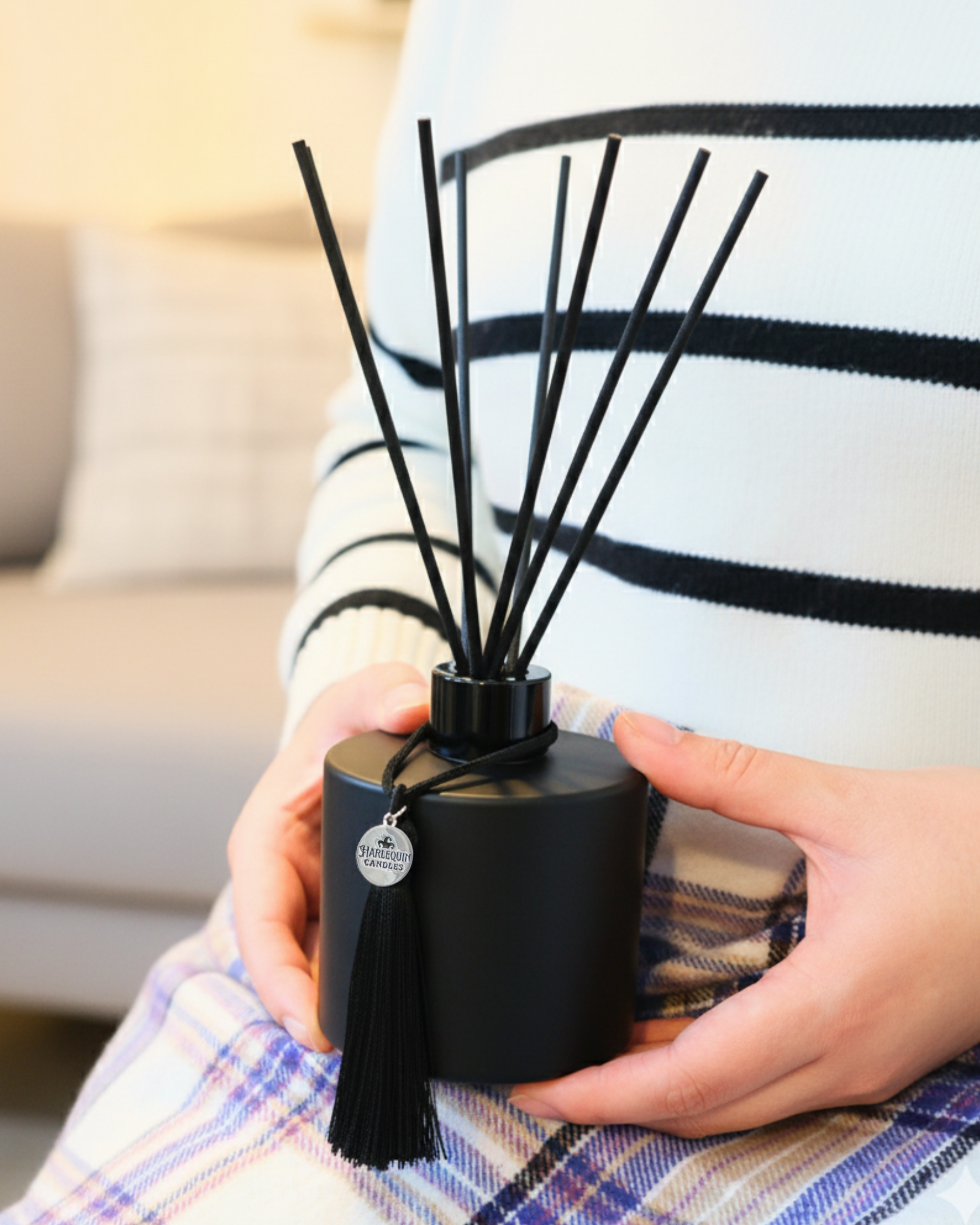Person holding a Harlequin Candles black reed diffuser with tassel in a cozy, warm living space. Premium home fragrance for relaxation, décor, and aromatherapy.