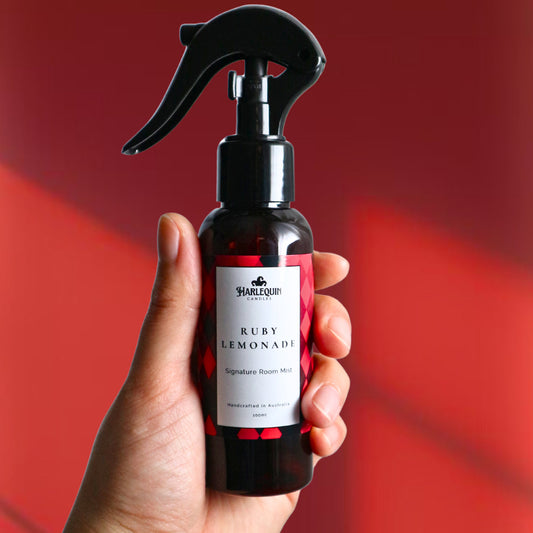 A Harlequin Candles Ruby Lemonade Signature Room Mist in an amber spray bottle with a black trigger cap, placed against a two-toned background of light pink and white. The label clearly shows 'Harlequin Candles', 'Ruby Lemonade', 'Signature Room Mist', and 'Handcrafted in Australia', highlighting this vibrant Australian-made home spray.