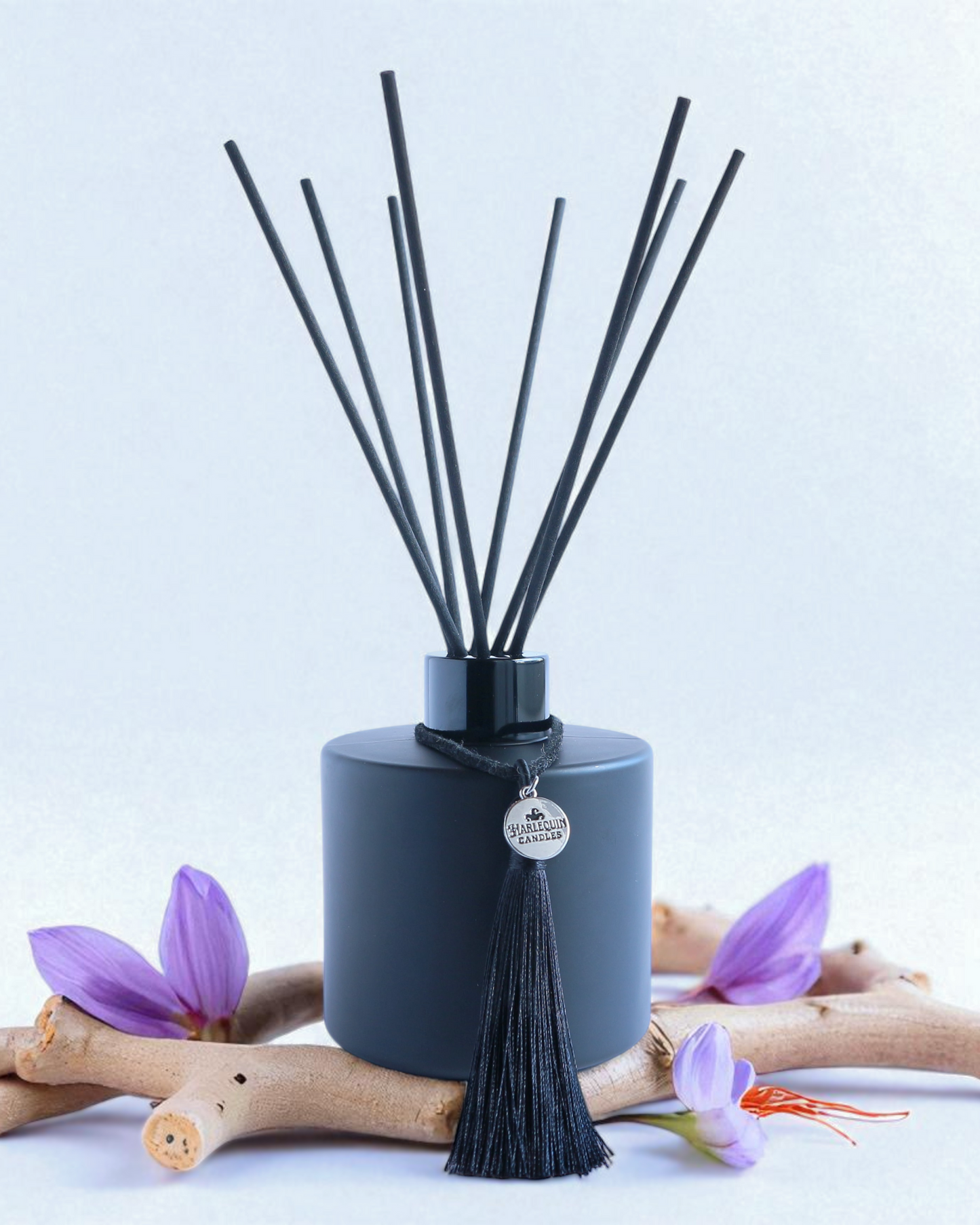 Harlequin Candles Saffron & Sandalwood reed diffuser in a matte black bottle with black reeds and a decorative black tassel with a silver logo charm. It is set against a blurred background of warm red and beige tones, evoking a rich and inviting home fragrance.