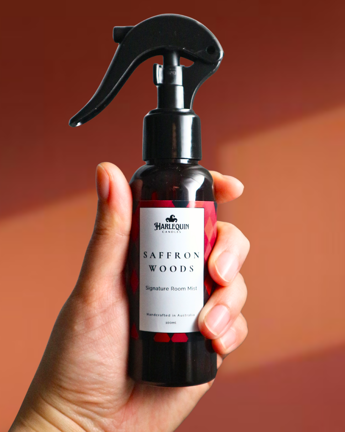 A Harlequin Candles Saffron and Sandalwood Signature Room Mist in an amber spray bottle with a black trigger cap, placed against a two-toned background orange and brown. The label clearly shows 'Harlequin Candles', 'Saffron Woods', 'Signature Room Mist', and 'Handcrafted in Australia', highlighting this enchanting Australian-made home spray.